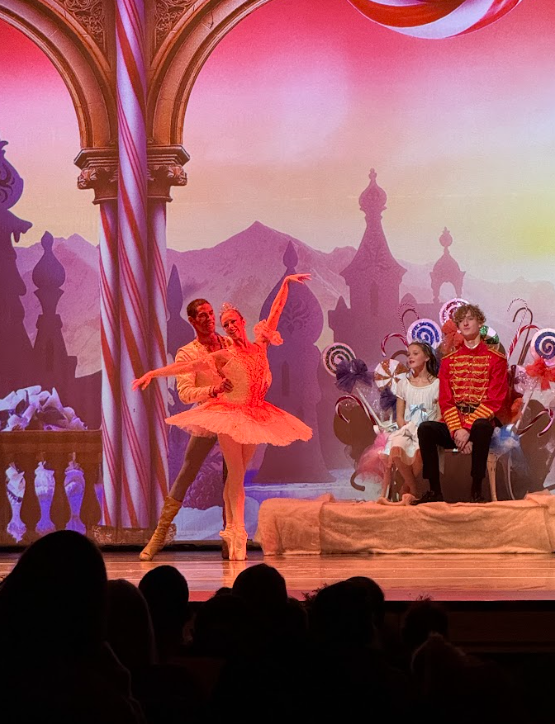 Sugar Plum Fairy ballerina in pink tutu with Cavalier pose together on stage while Clara and the prince watch in the Land of the Sweets background