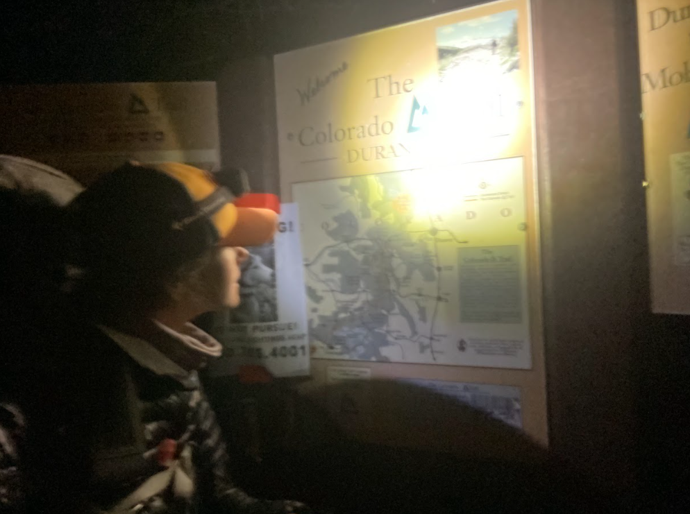 blurry image of a hiker in hat and headlamp looking at a trailhead billboard map of the Colorado Trail