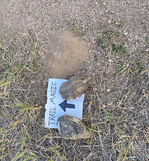 rocks pin down a handwritten sign on ground that reads "TRAIL MAGIC" with an arrow