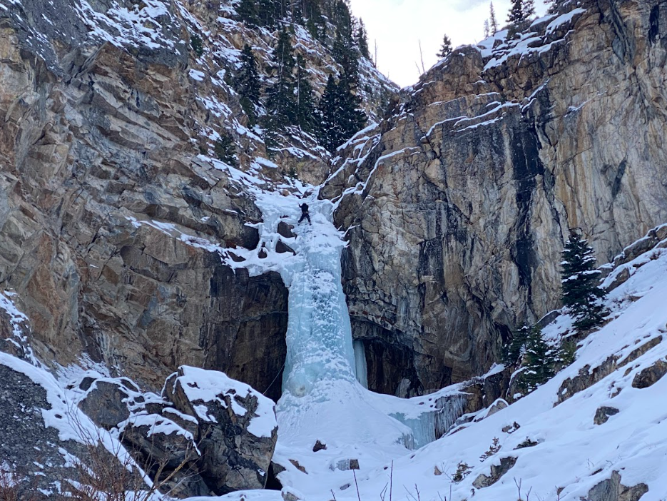 climber in black stands on ice from frozen waterfall between rocky outcroppings
