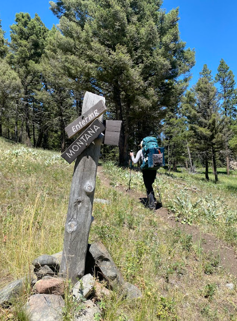 rickety wood sign states "Entering Montana" while a hiker continues on trail past it towards large trees