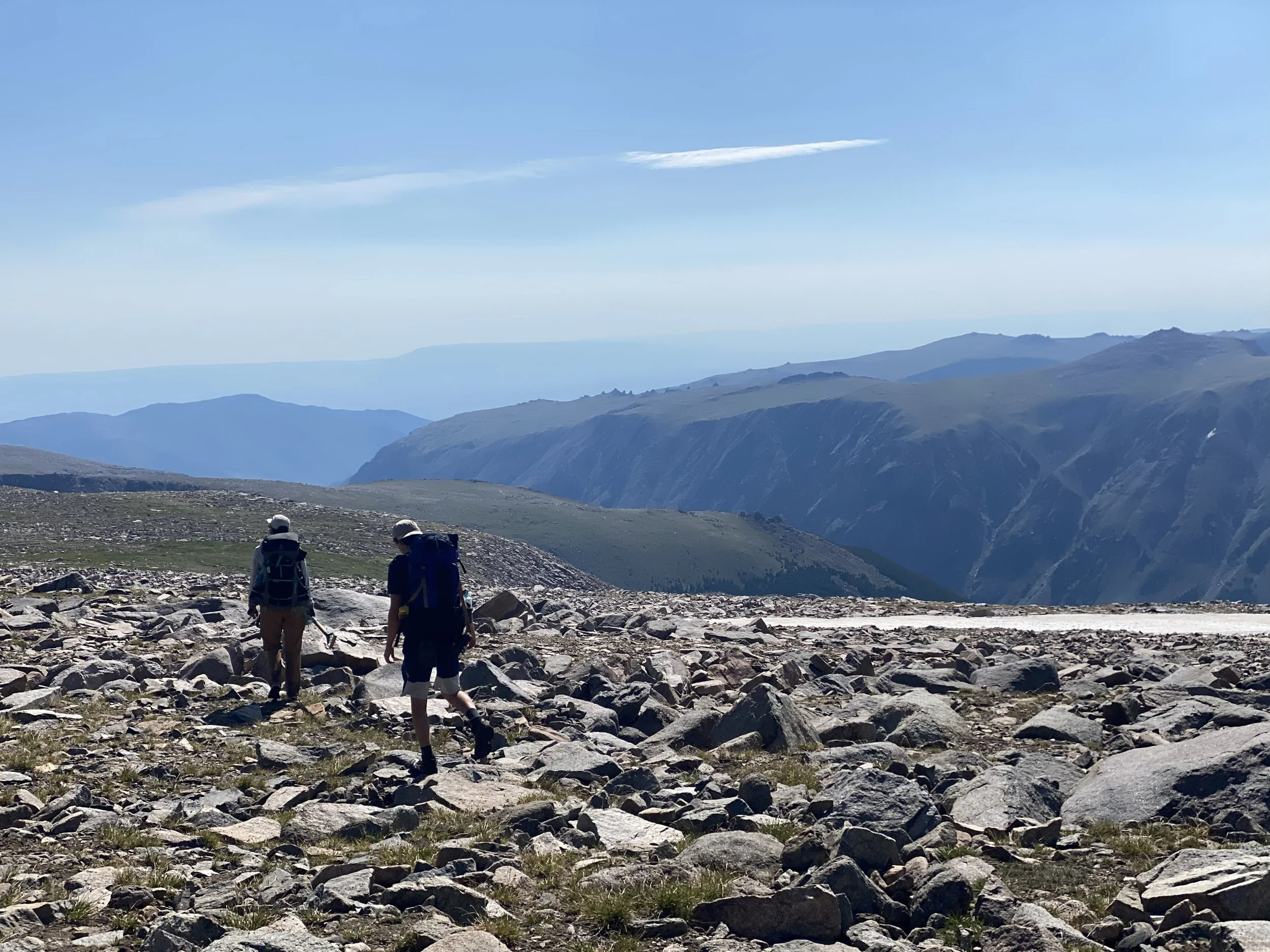 two hikers cross a boulder field along the edge of a plateau