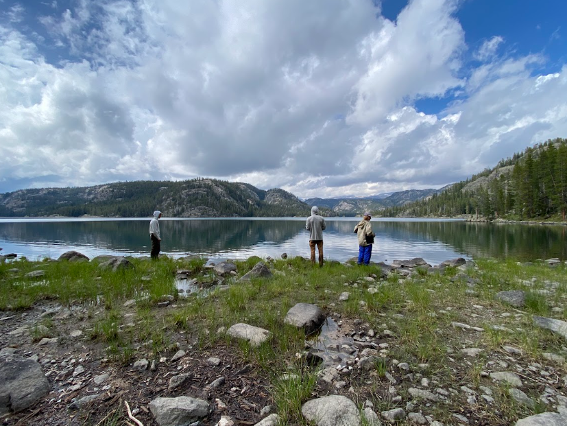 3 people stand on a lakeshore admiring the expanse of water under a cloudy sky