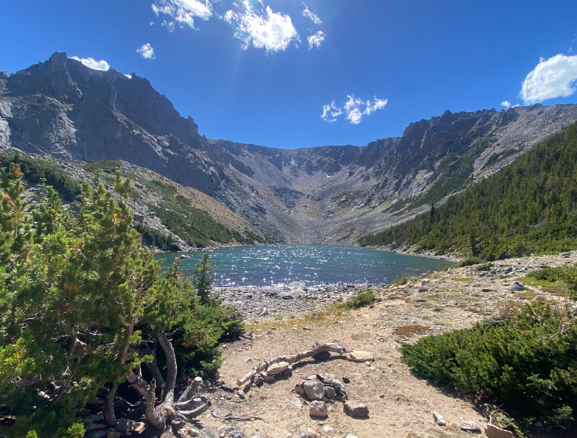 a small lake nestled under mountains with a sandy beach and shrub pines