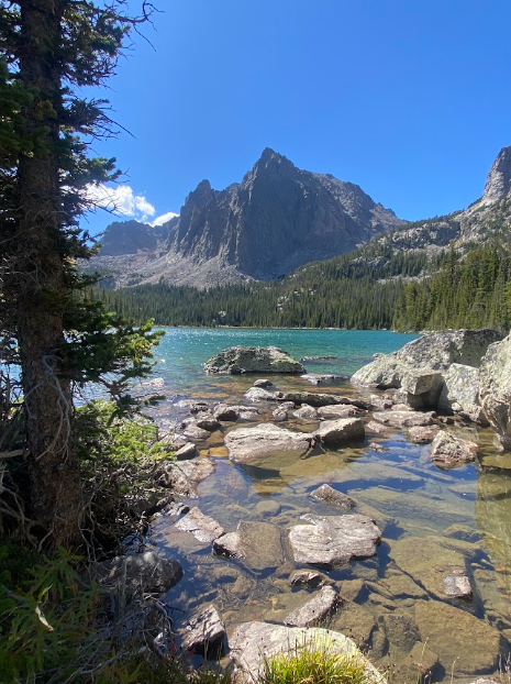 large rocks lead to a jewel toned lake that sits beneath a sharp triangular shaped mountain under a clear blue sky
