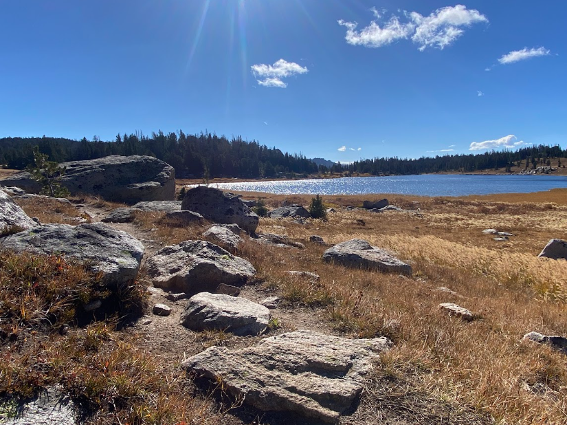 rocks along a trail skirt a blue lake under a clear sky