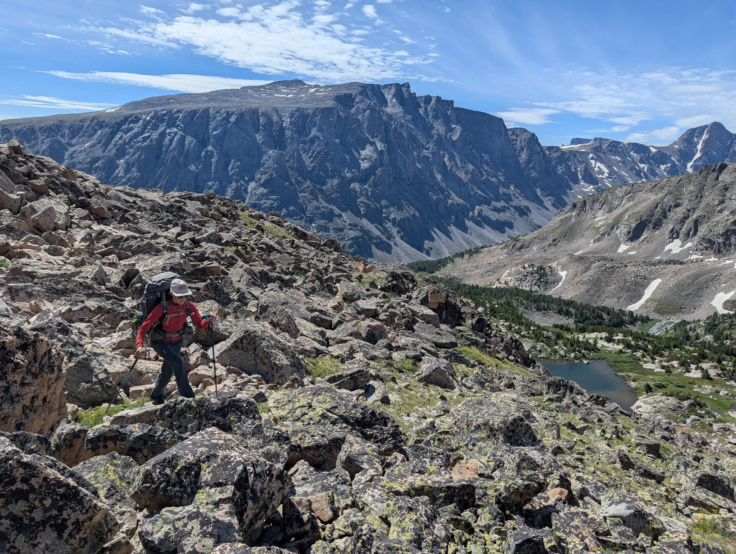 hiker in red shirt and large backpack crosses large rocks while the view of a lake spreads out behind her and massive rock walls of granite are in far distance