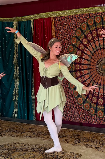 a dancer in green fairy costume with brown belt and golden wings poses with one arm lifted and looks down toward arm that is lowered