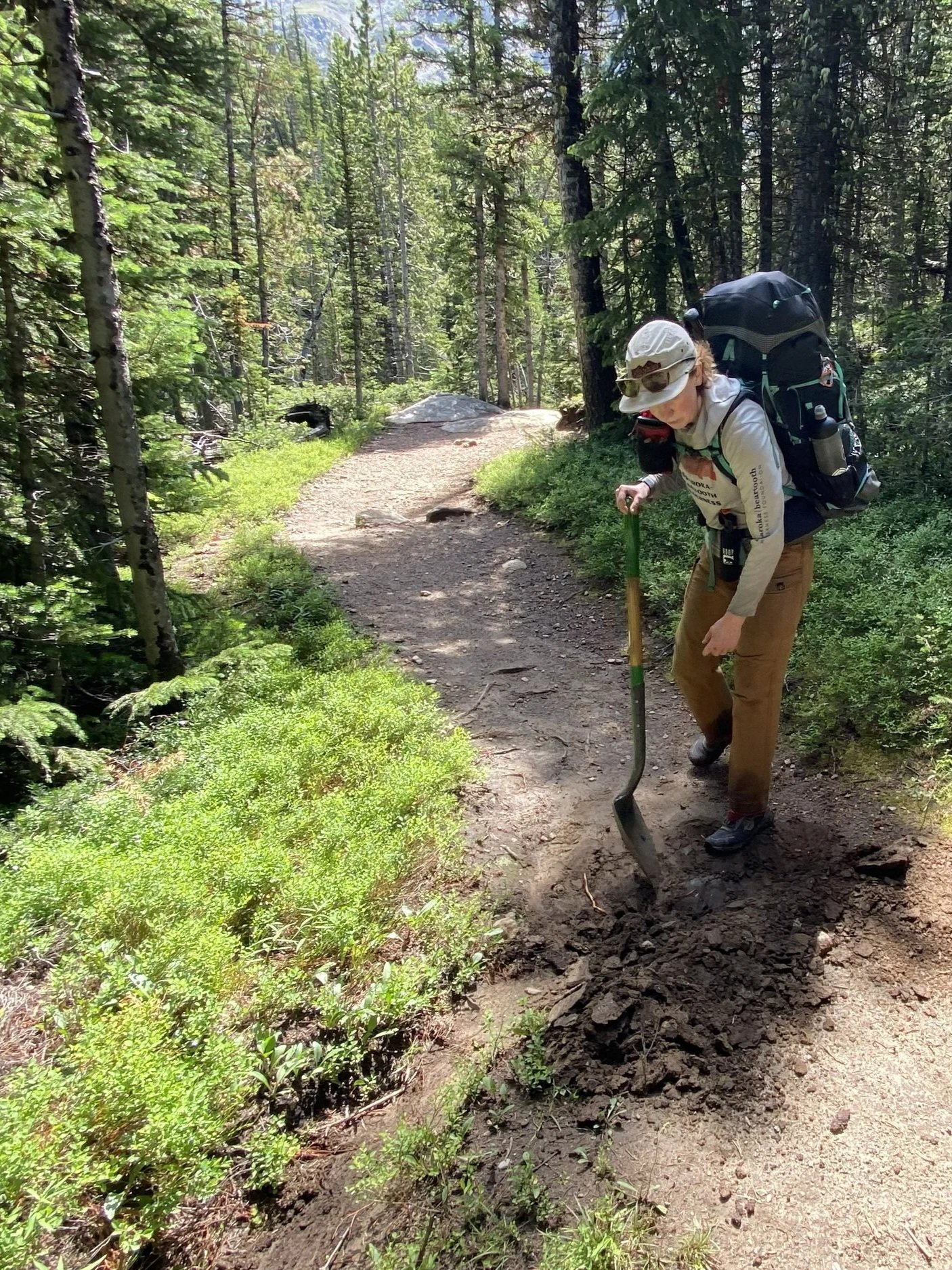 a hiker holds a shovel and points over broken up dirt while on a trail in the woods