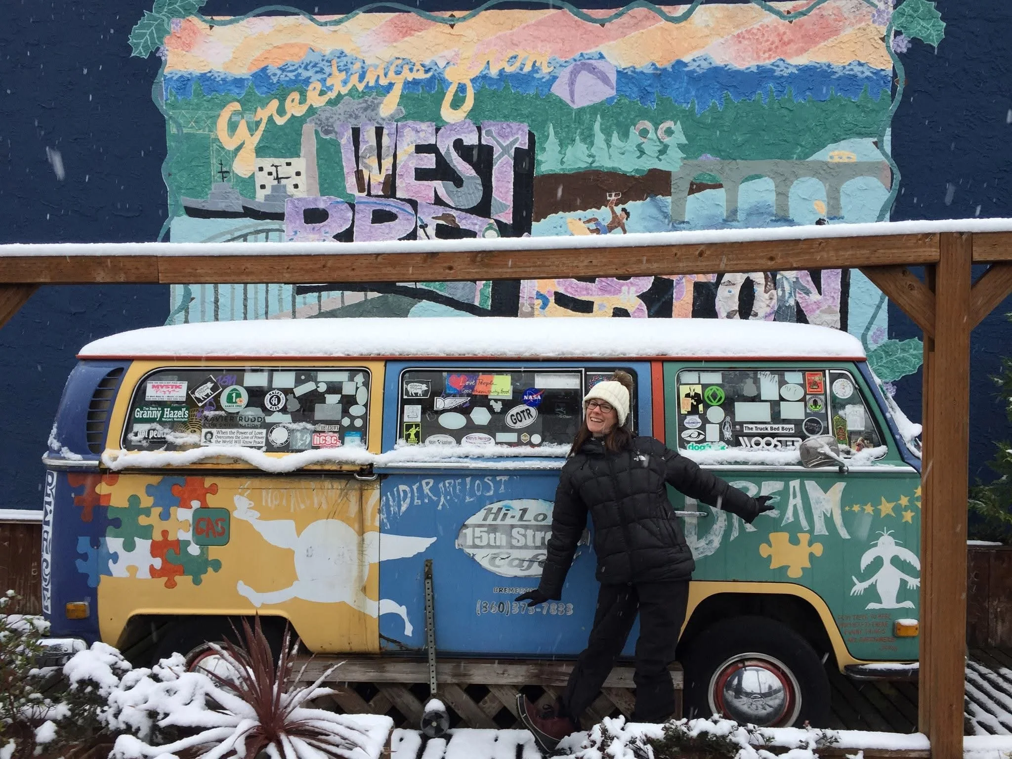woman in black winter clothes poses in front of VW van that is painted with bright colors and sits in front of a West Bremerton mural