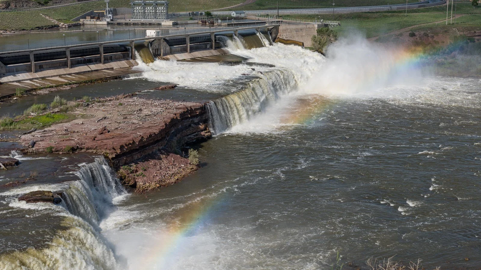 a dam with rocky outcropping beneath it leading to a wide, short waterfall across a murky river