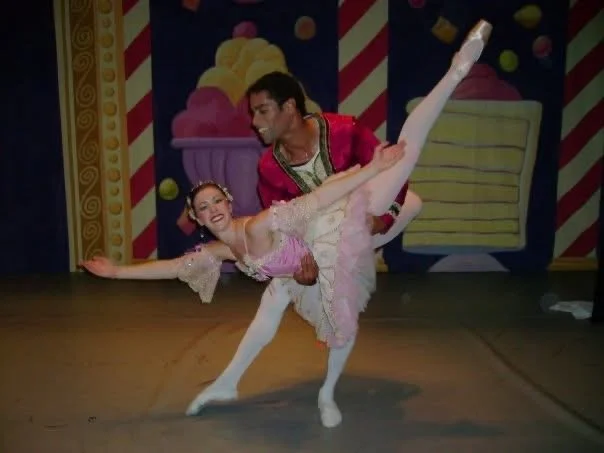 the author smiles at the camera in pink tutu is held low in the air by her partner, a black man in red jacket and white tights
