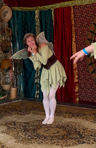 female dancer in green fairy costume with wide brown belt and golden wings acts frightened and backdrop includes bright tapestry