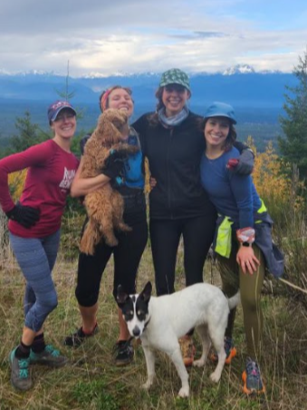 four ladies wearing running clothes pose with two dogs in front of a panoramic view of mountains