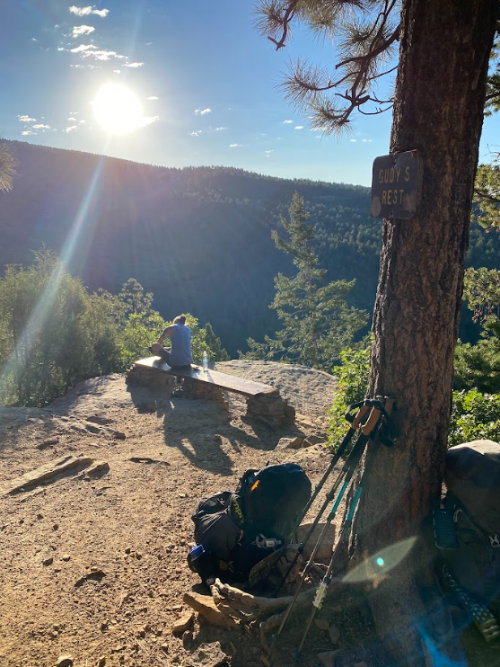 backpacks and poles rest against a pine tree that has a sign stating Gudy's Rest while a man sits on a bench enjoying the view and bright sun