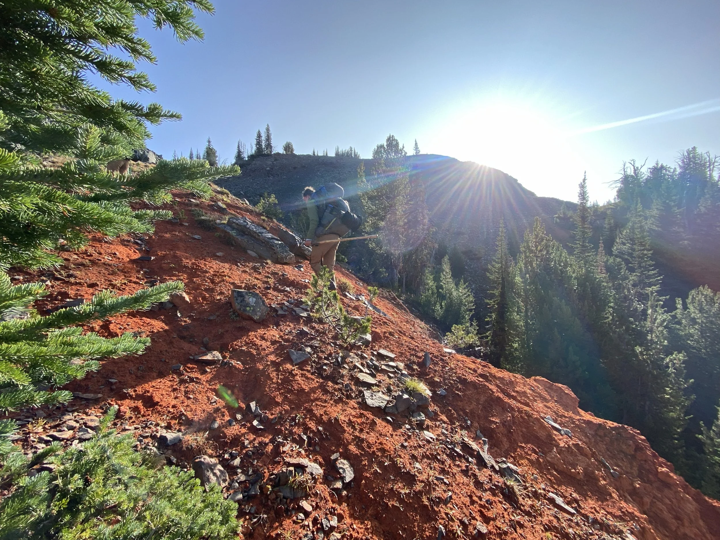 a man walks up a hillside of bright red soil while the sun rises over the distant mountain peak