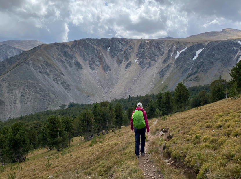 A hiker dressed in pants and pink rain jacket walks along narrow trail on a steep hillside towards a view of a crater like bowl of granite mountain with a dark sky above
