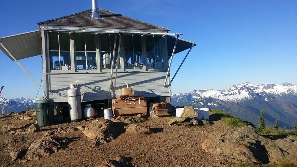 gray squat fire lookout tower on a ridgeline of dirt with a snow covered mountain range close in the distance