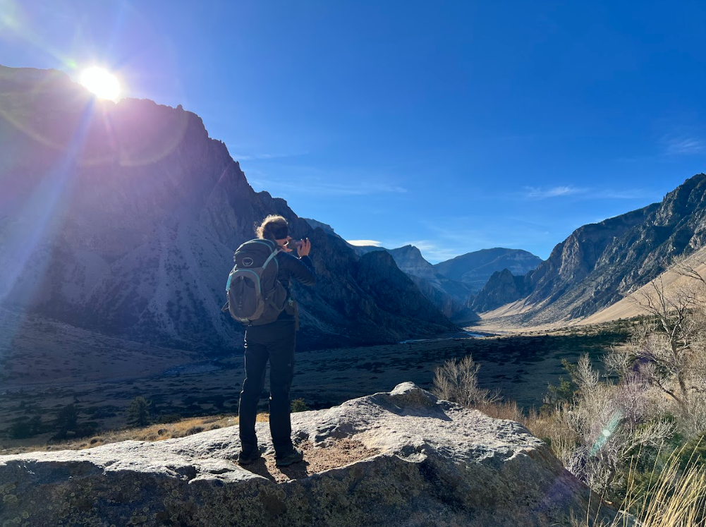 hiker taking a photo of a canyon on a bluebird day while the sun shines brightly over the left mountain peaks