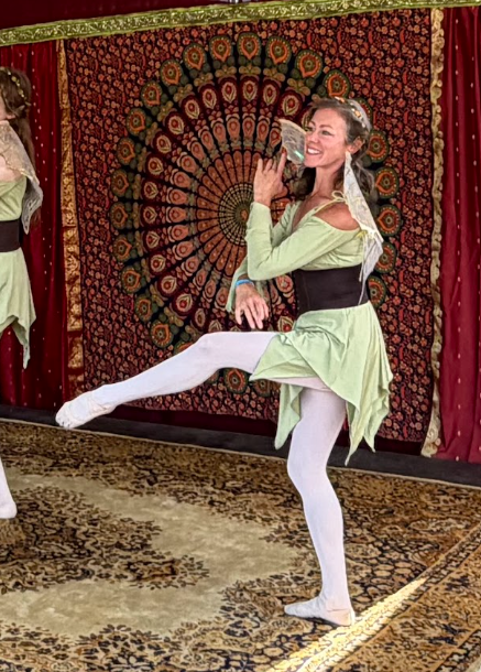 dancer in green fairy costume with golden wings and wide brown belt balances on one leg while extending the other leg in front