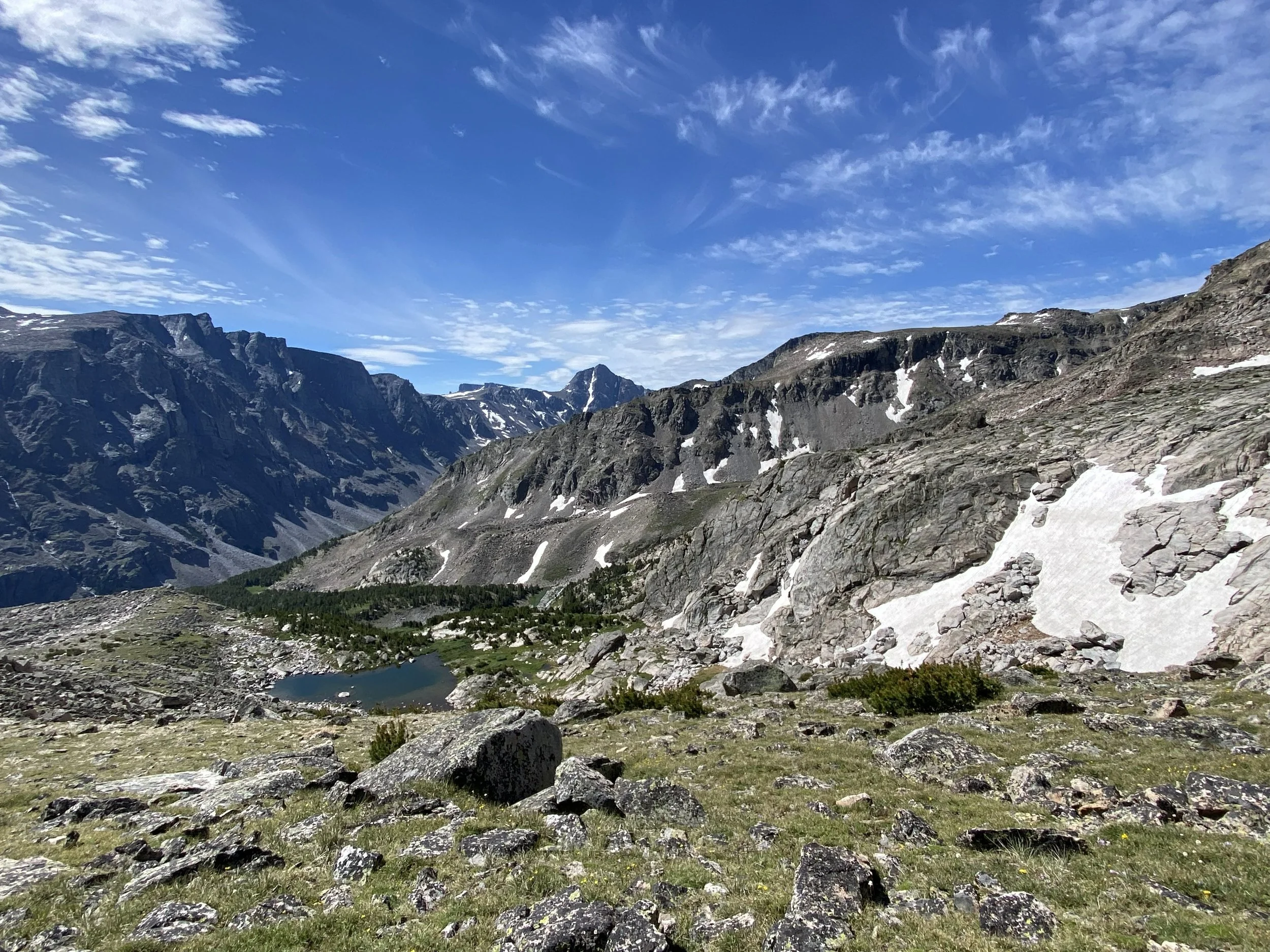 overlooking a lake with a backdrop of granite rock outcroppings holding patches of snow, grass, and trees under a bright blue sky