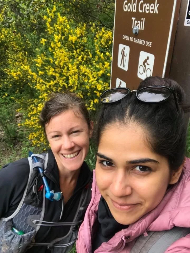 two women in hiking gear smile for selfie in front of the Gold Creek Trail sign and yellow flowering bushes