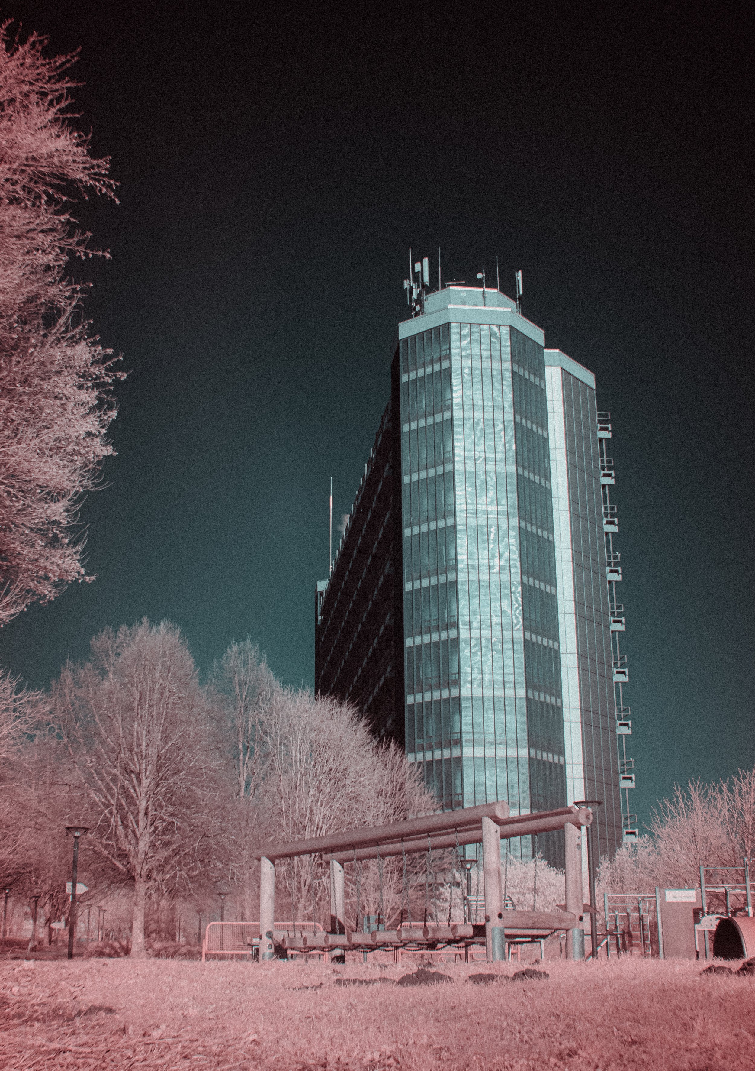 Infrared photo of a tall glass building with antennas on the roof, surrounded by leafless trees and a park area with playground equipment in the foreground.
