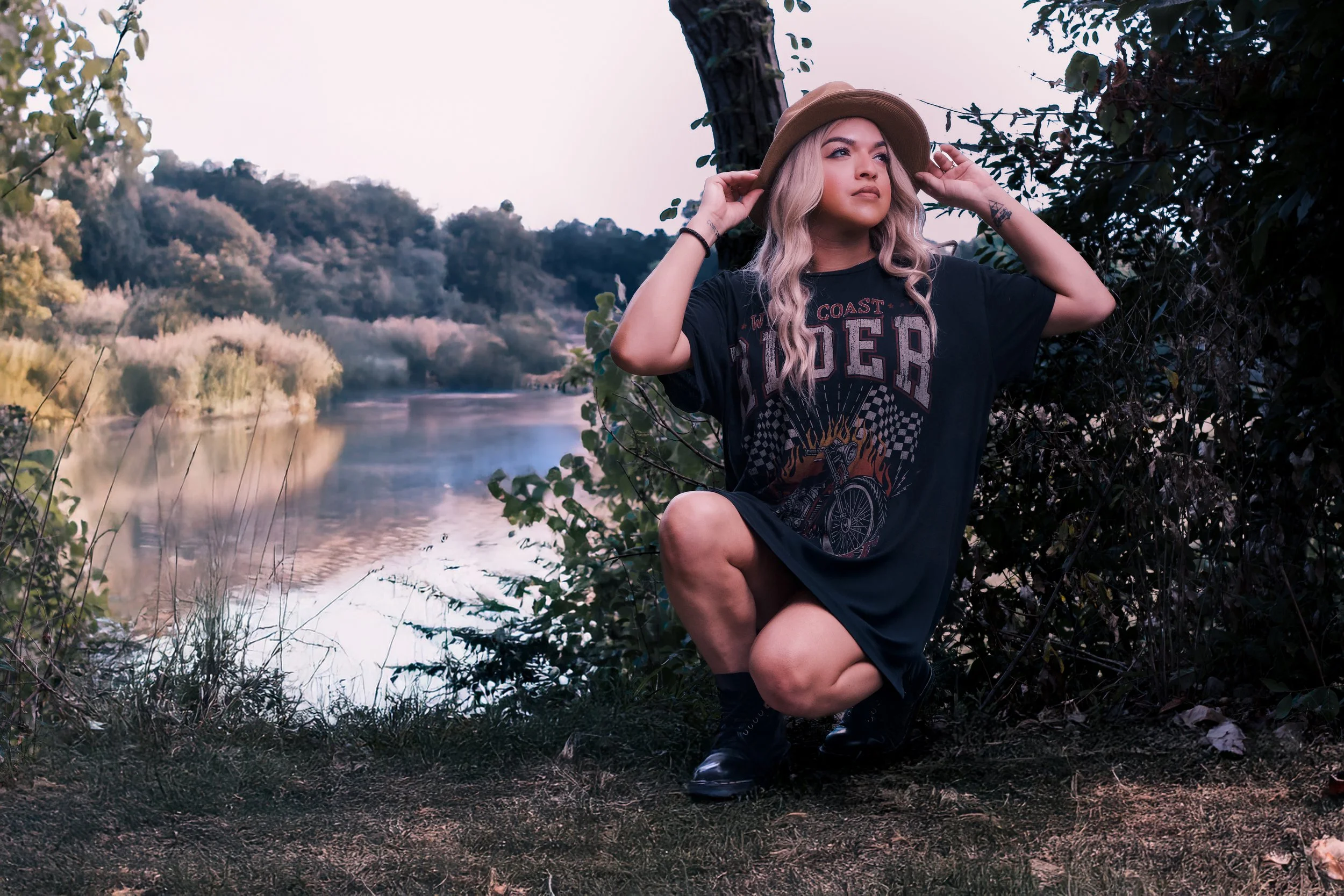 A woman with long blonde hair in a beige hat, wearing a black graphic t-shirt, squatting near a river, surrounded by trees and bushes, with a wooded landscape in the background.