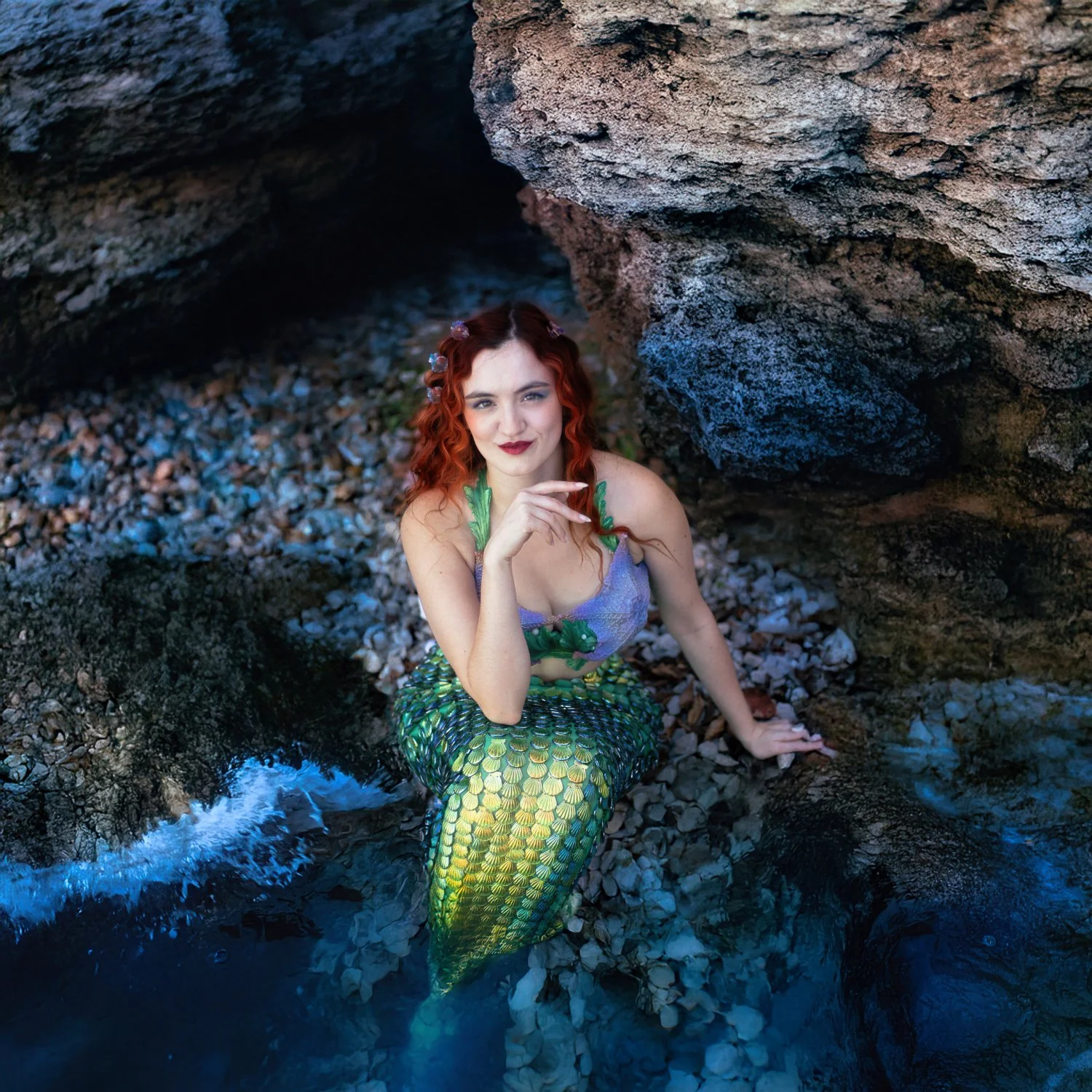A woman dressed as a mermaid, with long curly red hair and a purple top with green leaf accents, sitting in a rocky shoreline cove surrounded by water and large rocks.