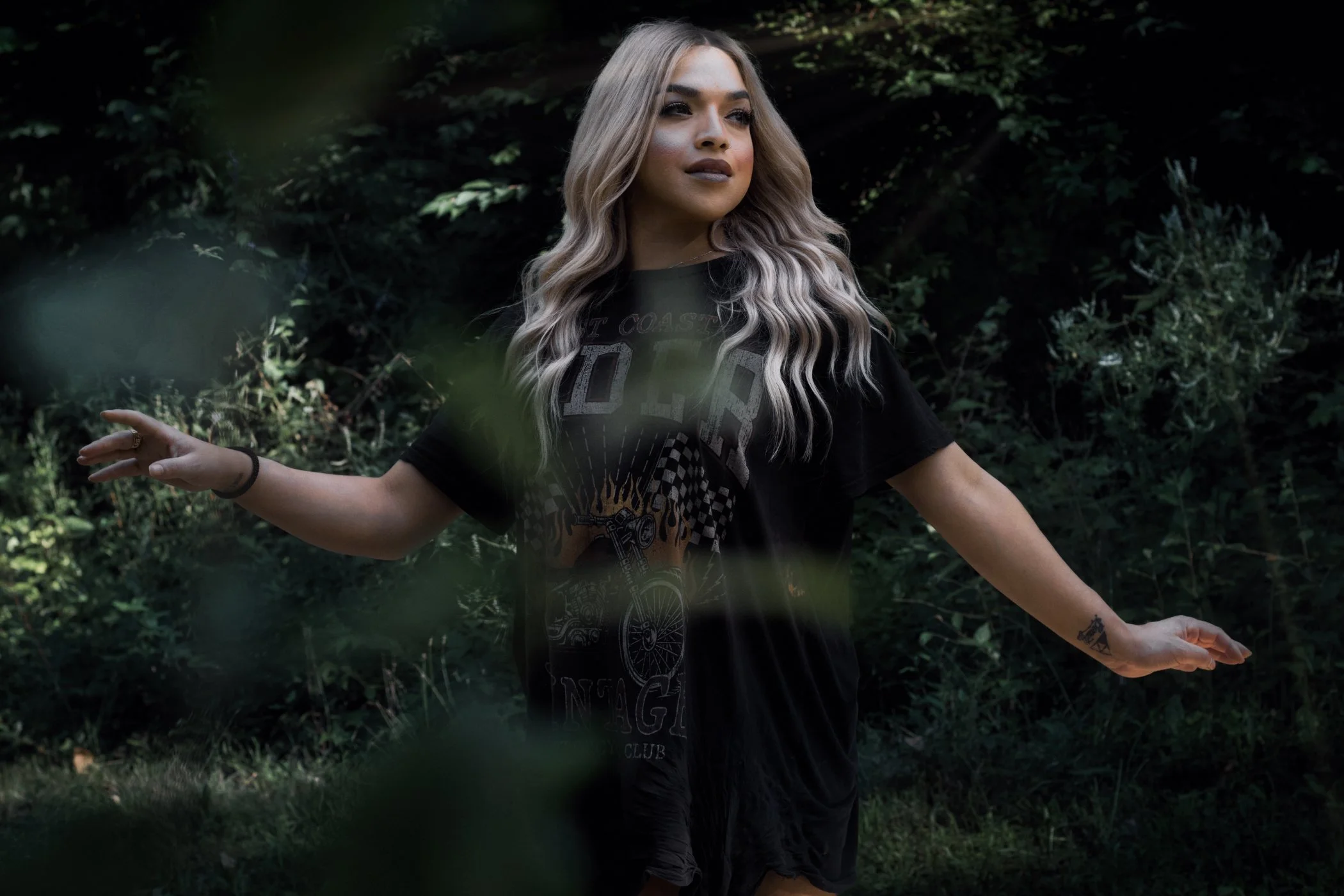 Young woman with long blonde wavy hair standing outdoors in a wooded area, wearing a black T-shirt with graphics, arms outstretched, and a tattoo on her right forearm.