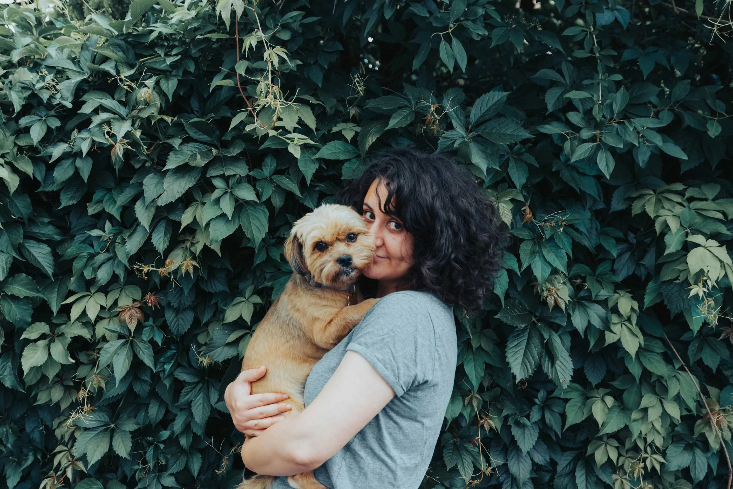 Woman with curly dark hair holding a small tan puppy with floppy ears against a background of dense green foliage.