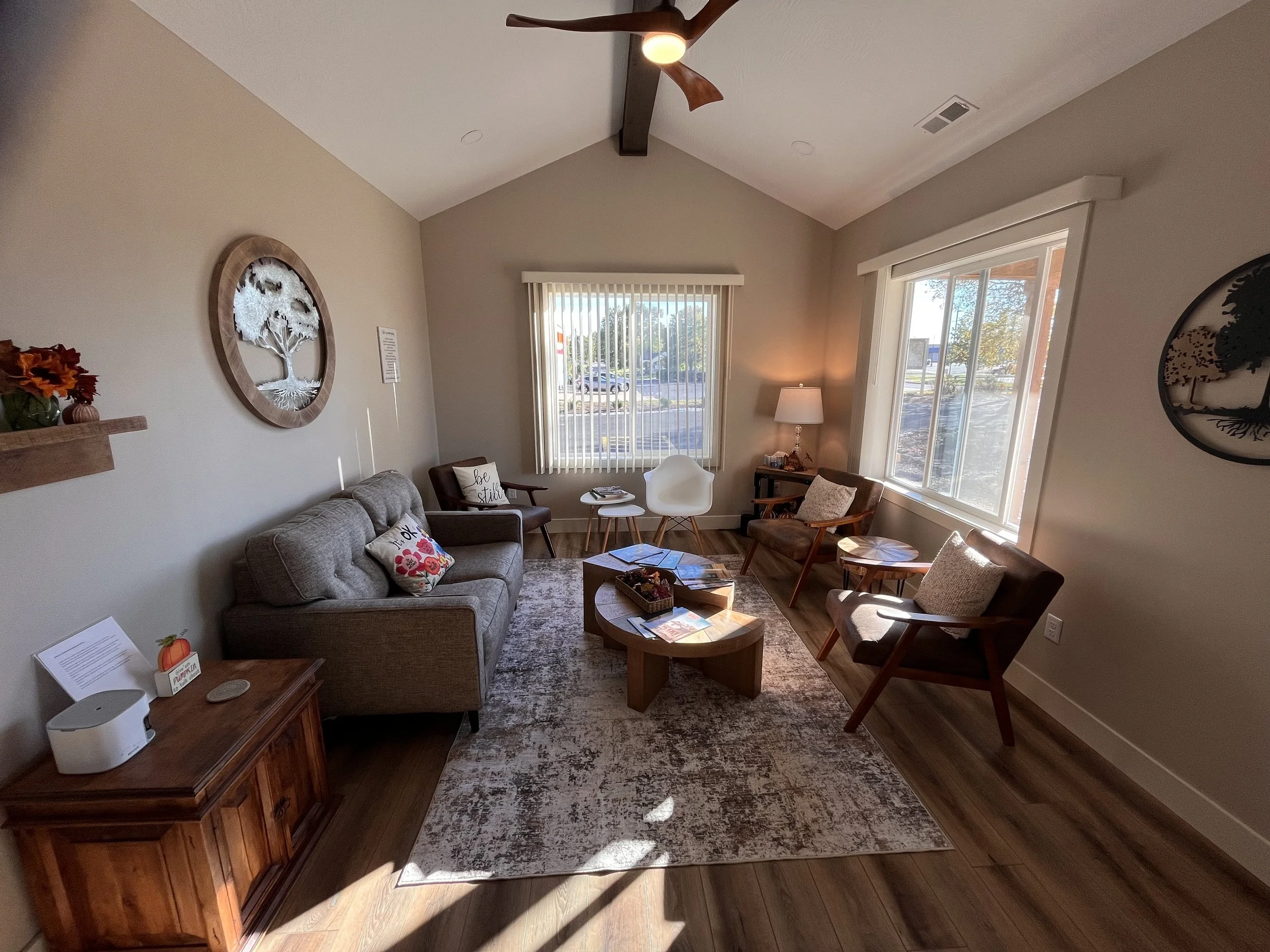 two wooden armchairs with beige cushions, a white modern chair, and a round wooden coffee table. There is a beige area rug, a wooden side table with a lamp, and large windows with vertical blinds allowing sunlight to fill the room. Decor includes a wall-mounted wooden tree art, a small shelf with fall flowers, and cushions with decorative pillows.