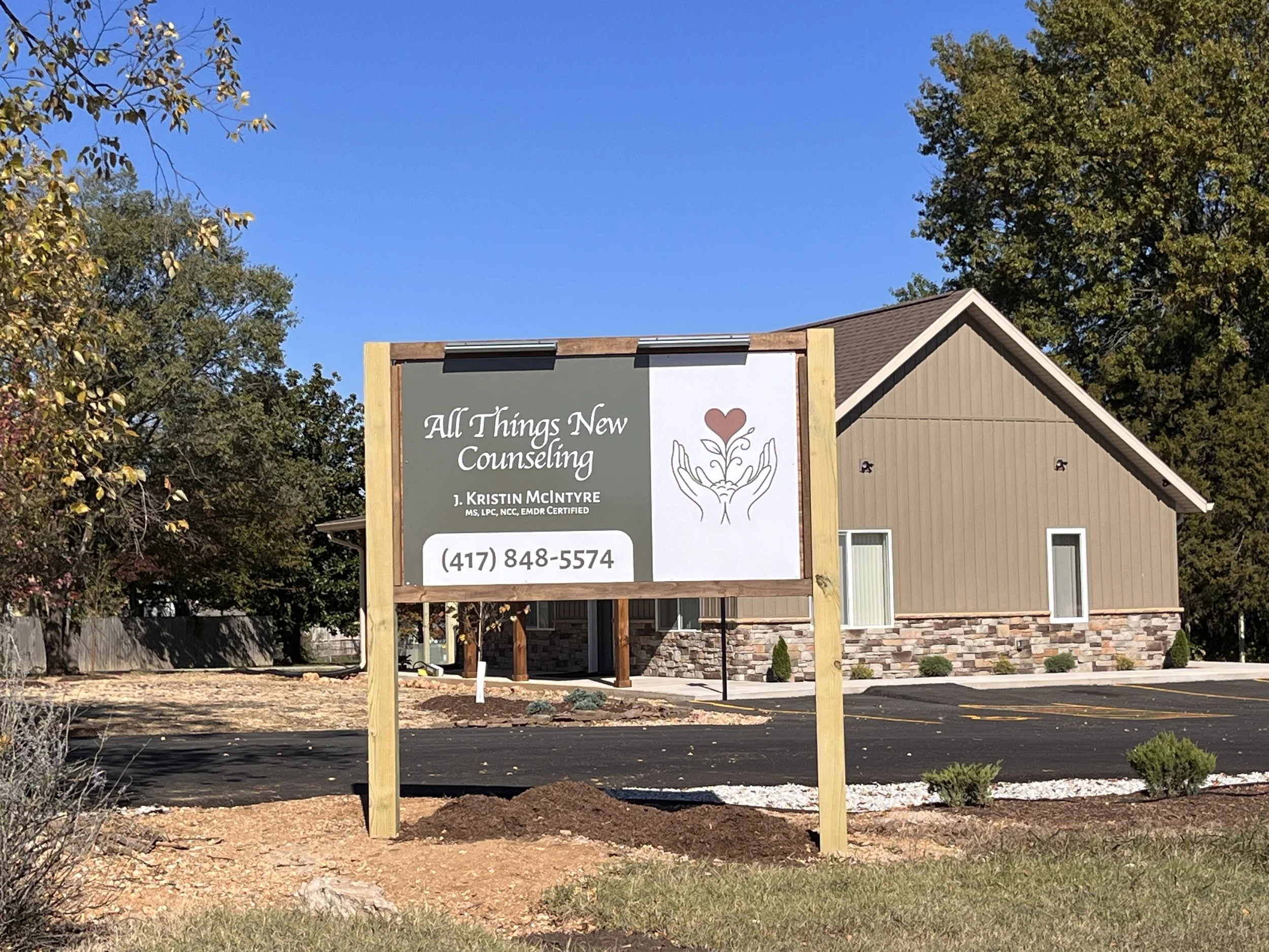 A sign for All Things New Counseling with contact information, located in front of a beige building with small windows, trees, and a clear blue sky in the background.