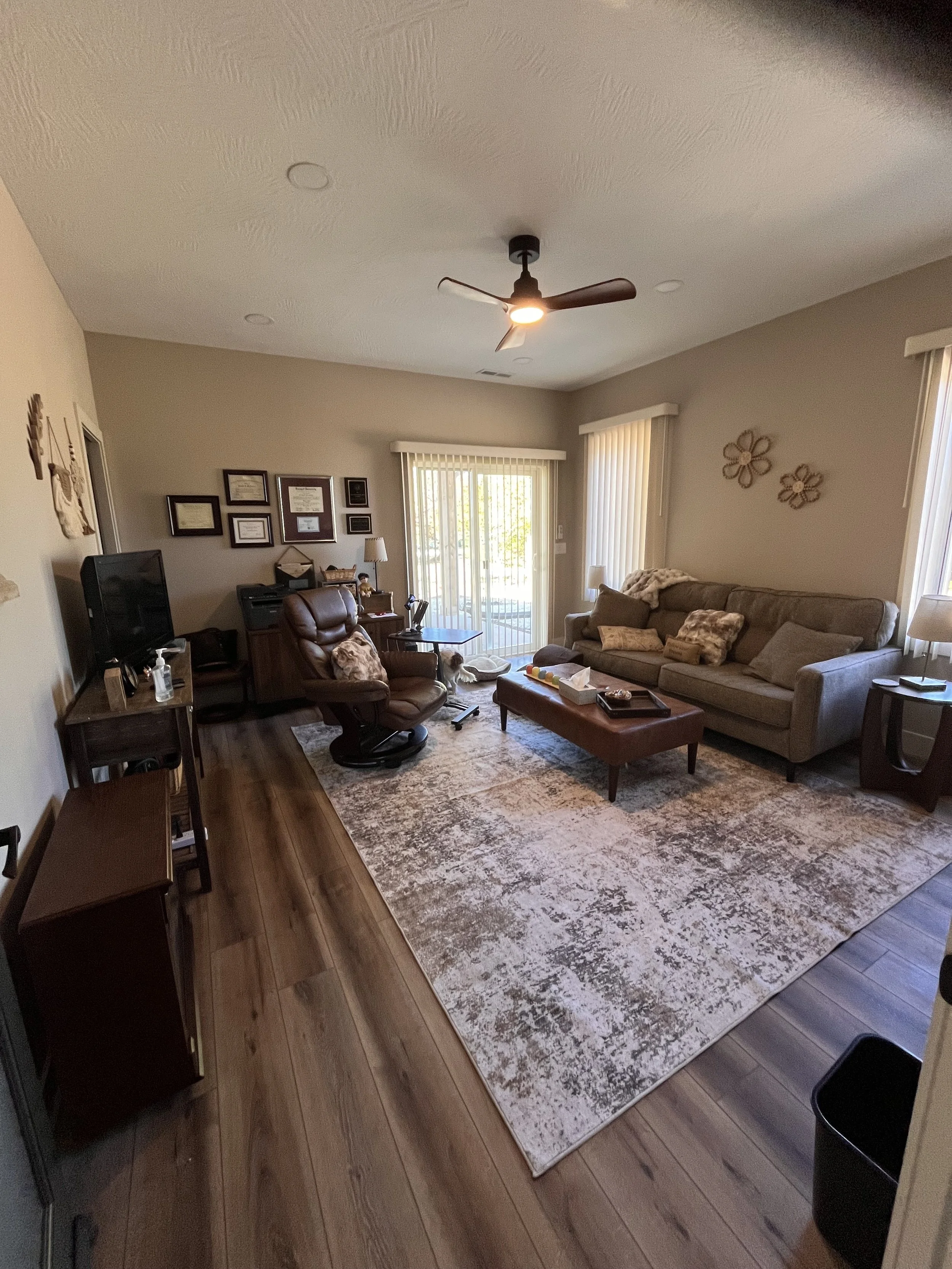 Kristin McIntyre's therapy room with a beige couch, brown leather chair, area rug, and various wall decorations. There is a sliding glass door with vertical blinds and a ceiling fan.