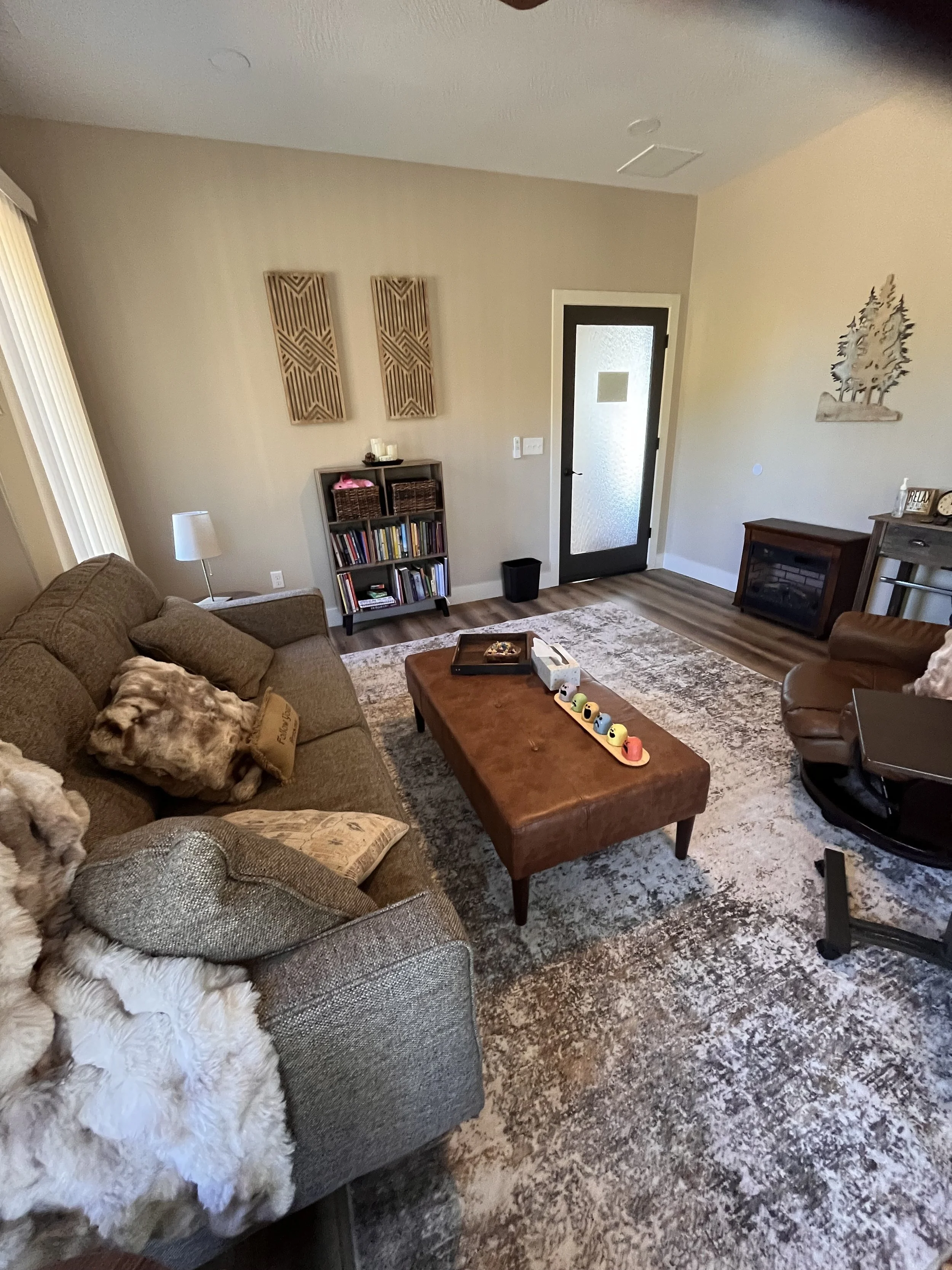 Kristin McIntyre's therapy room with beige walls, a tweed couch with soft fur and patterned pillows, a round chair, a brown leather ottoman with toys, a bookcase, a fireplace, and wall art. There is a door with frosted glass and window blinds.