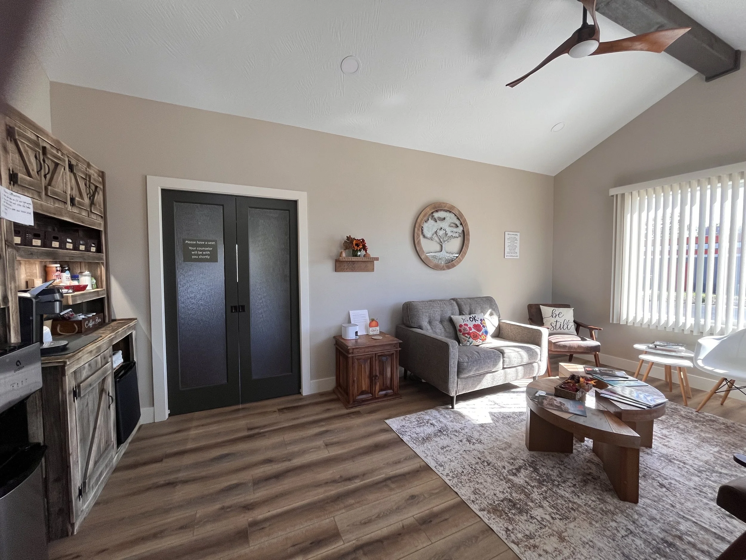 All Things New Counseling lobby with tweed sofa, wooden coffee table, armchair with pillow, window with vertical blinds, wooden shelf with coffee supplies, and wall decorations including a clock and floral arrangement.