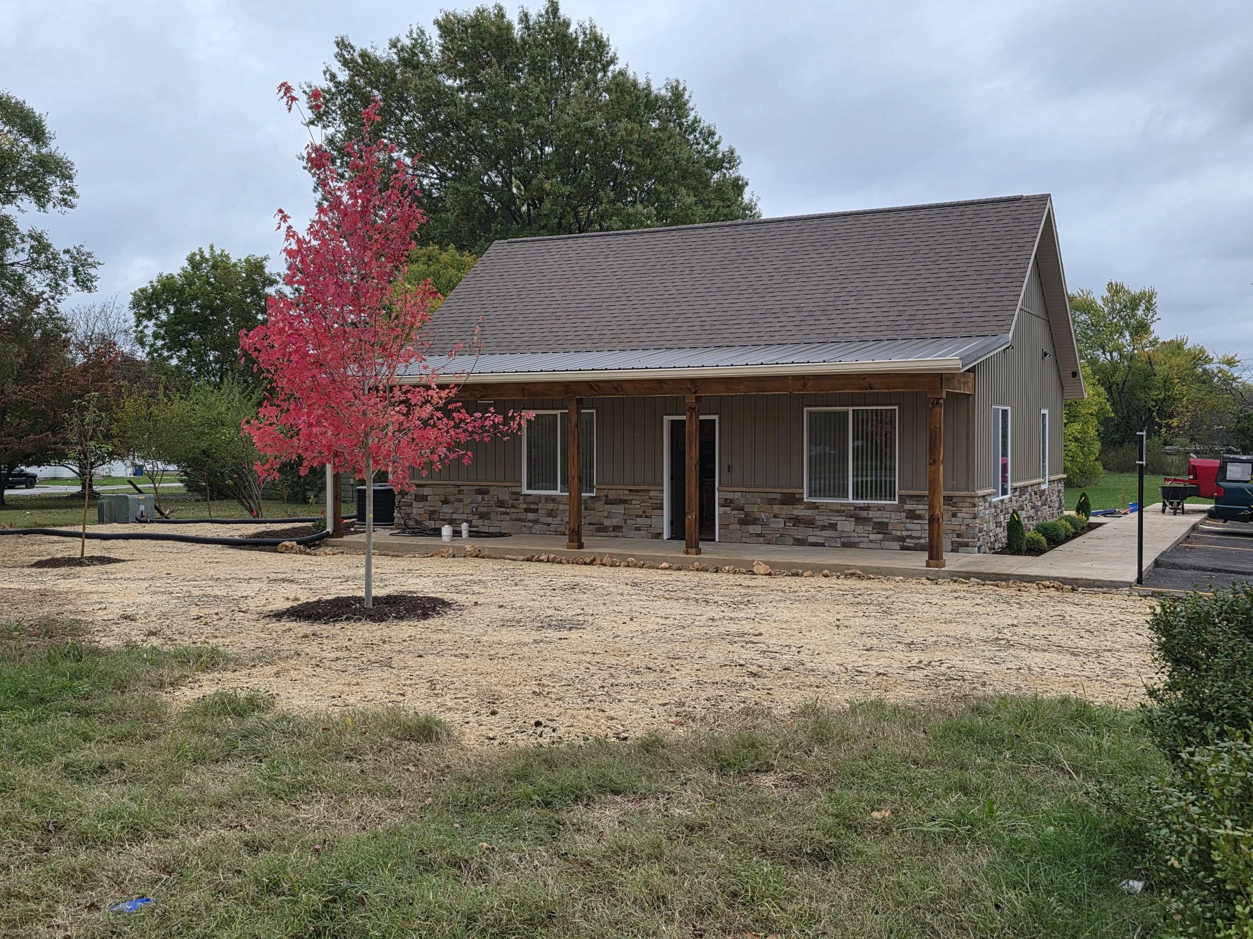 Newly constructed house with a porch, surrounded by trees, with a bright pink tree in front and a gravel area where grass has not yet grown.