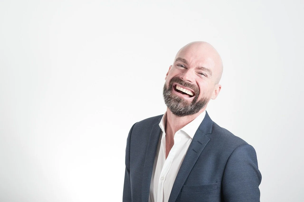 A bald man with a beard laughing, wearing a dark suit and white shirt, against a plain white background.