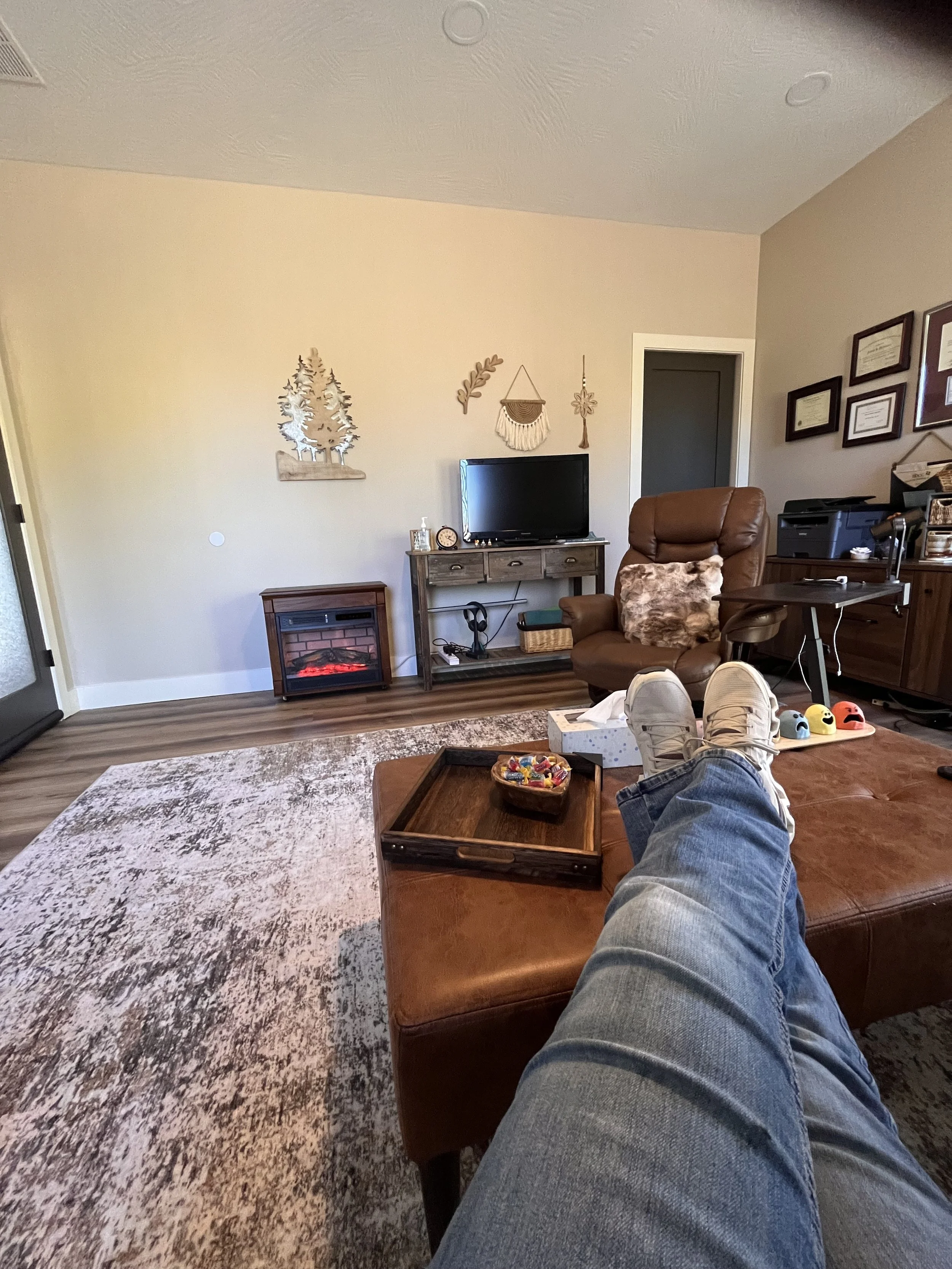 Feet up on a brown leather ottoman with legs crossed, wearing jeans and white sneakers, in a living room with a patterned rug, small wooden tray, TV on a rustic table, brown armchair, and wall decorations.