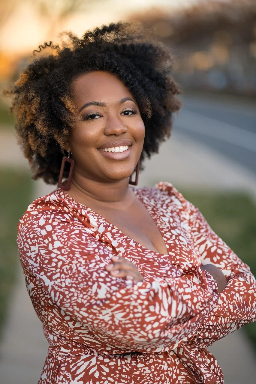 A woman with curly dark hair and hoop earrings smiling with arms crossed, wearing a patterned red and white dress, outdoors during the daytime.