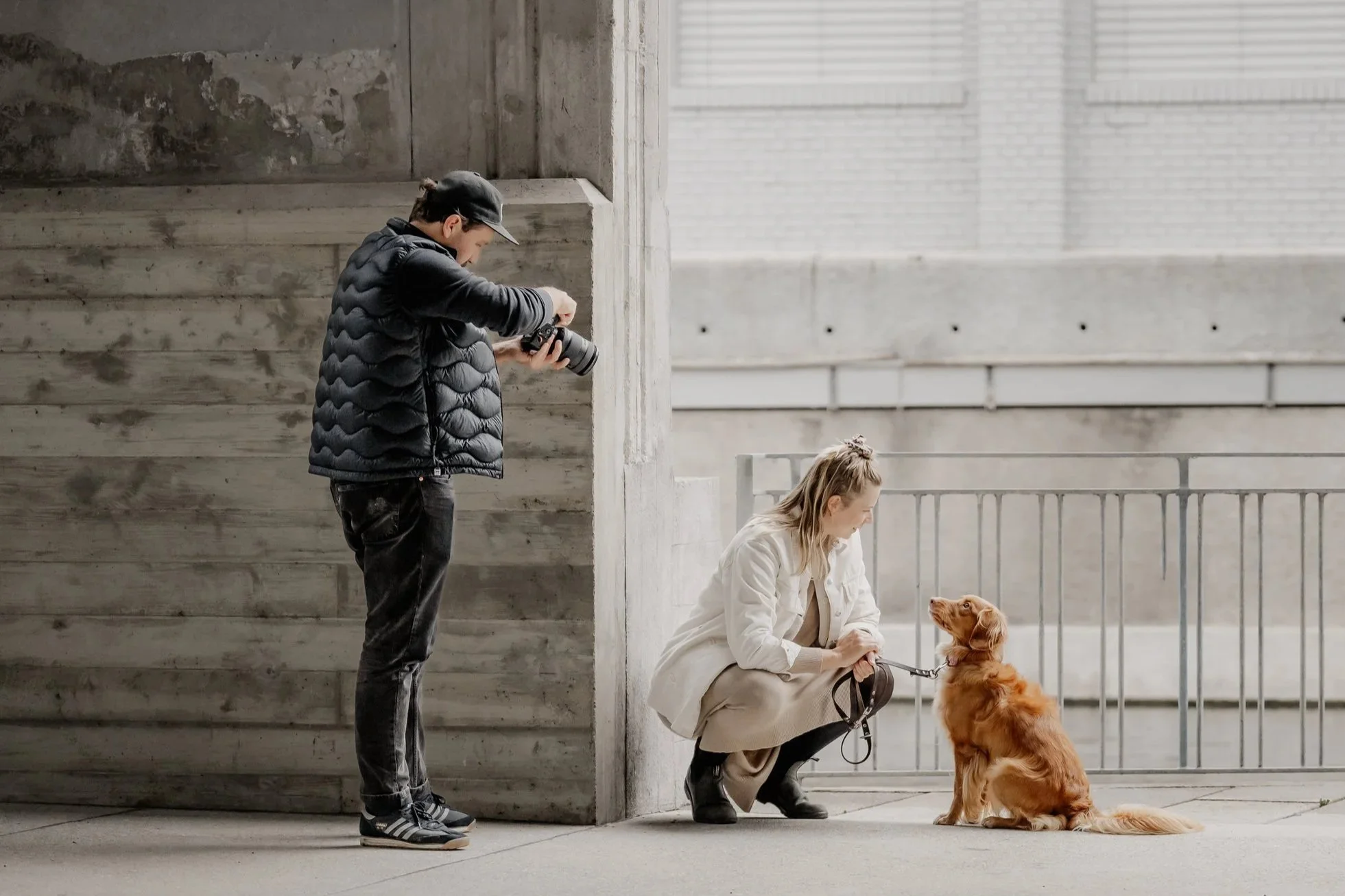 Ein Mann fotografiert eine Frau, die mit einem Golden Retriever Hund an einer Leine sitzt, in einer städtischen Umgebung mit Betonwänden und Metallzaun.