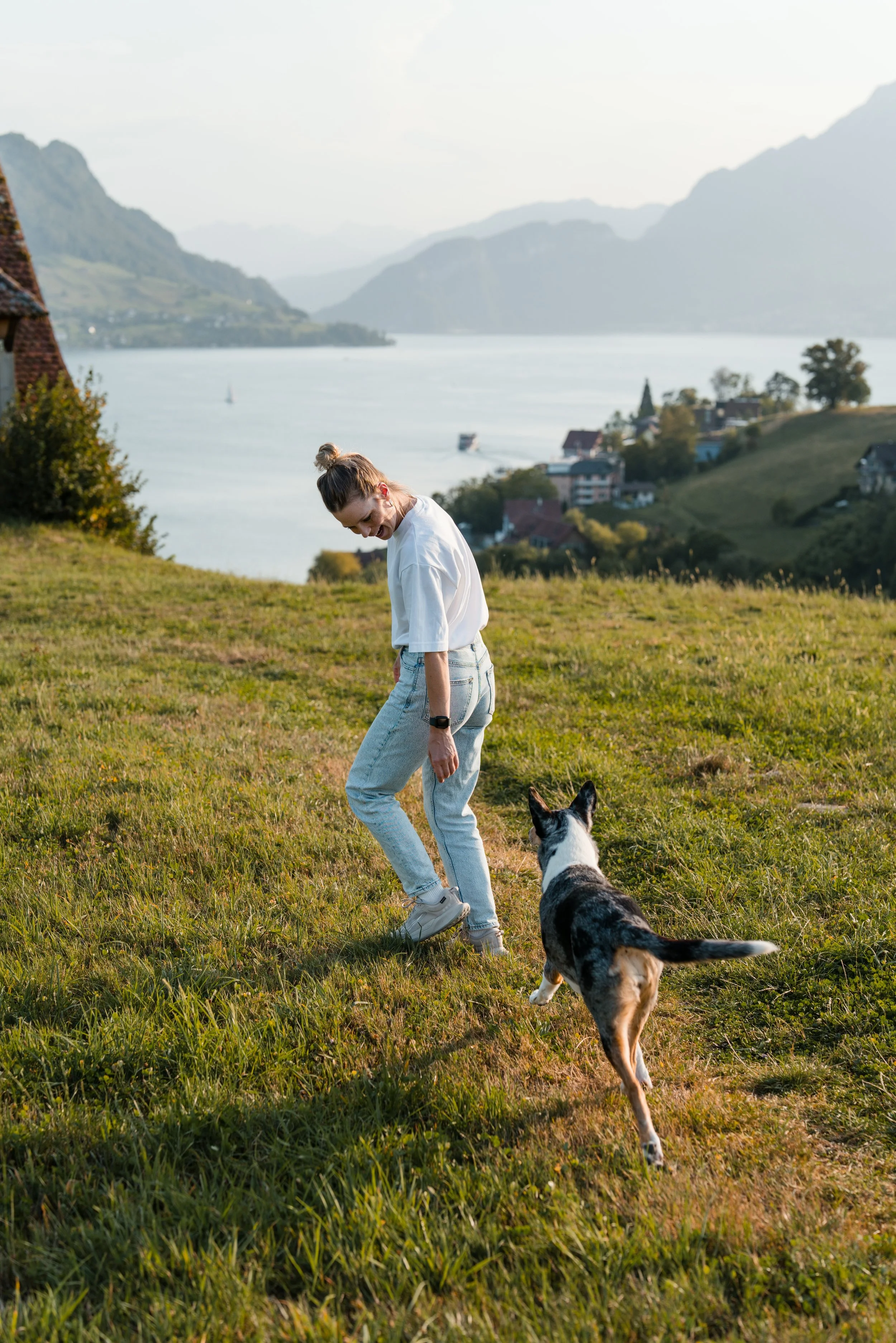 Frau spielt mit ihrem Hund auf einer grünen Wiese mit Blick auf einen See und Berge im Hintergrund in der Abenddämmerung.