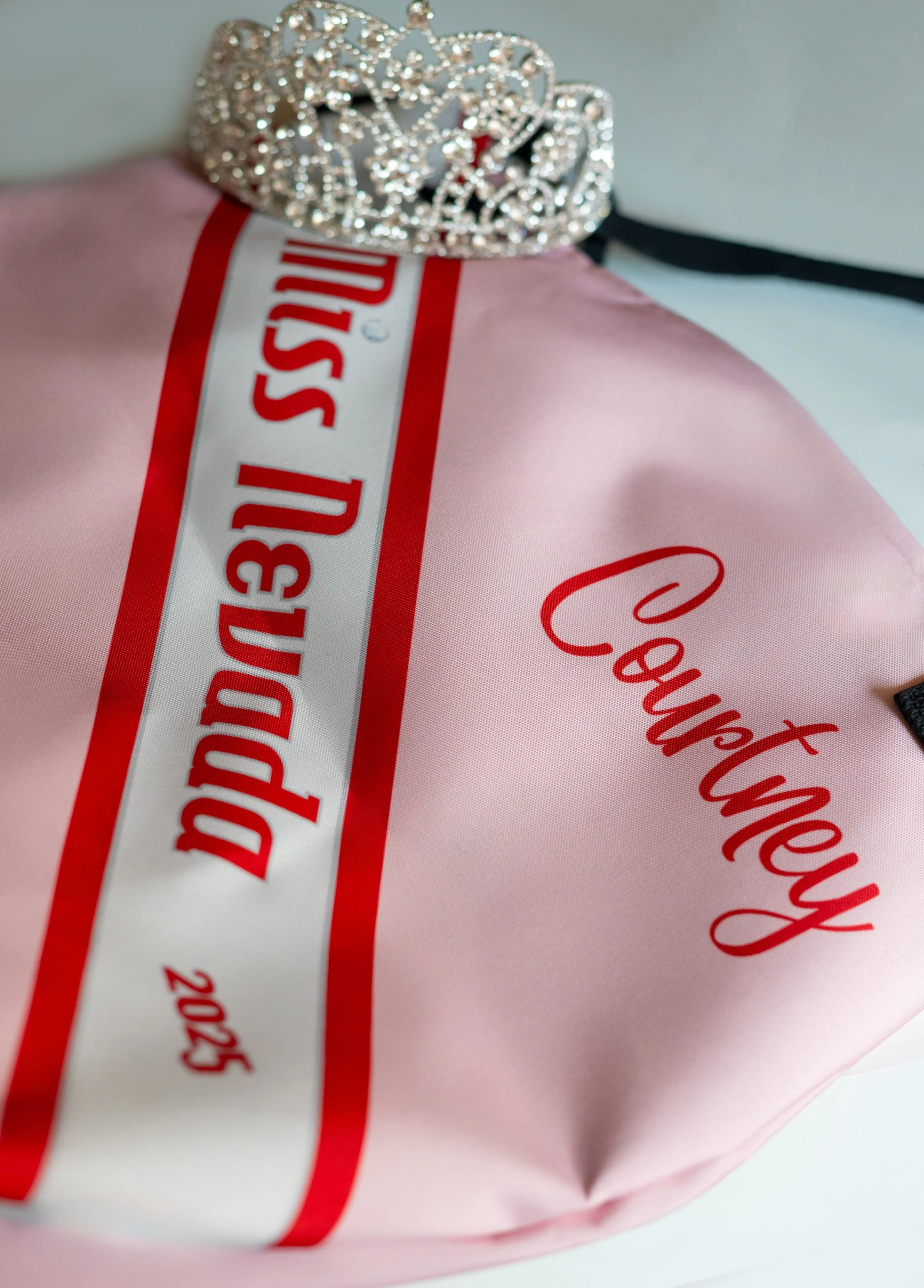 A pink satin pillow with the words 'Curtney' written in red cursive and a white label with red text that says 'DRESS UP' and '2025' with a silver rhinestone tiara on top.