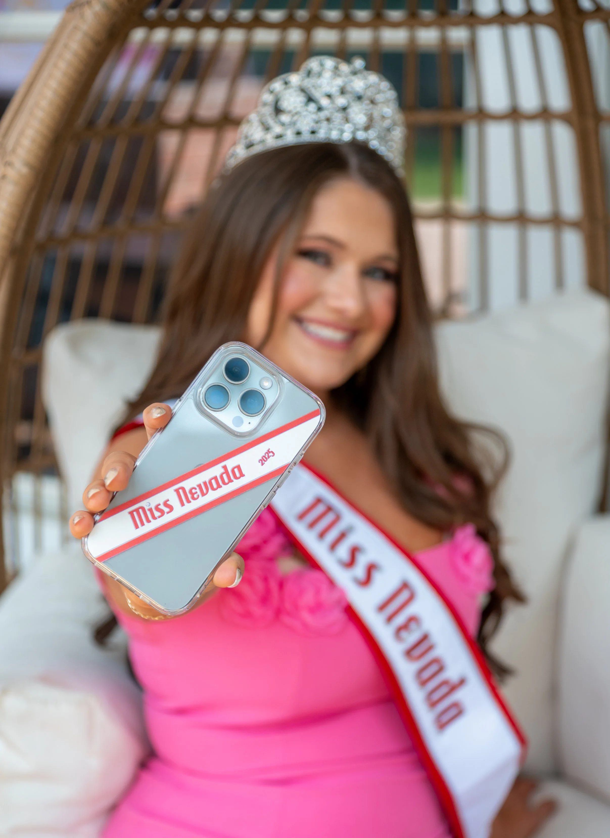 Miss Nevada 2025 wearing a pink dress and crown, smiling and taking a selfie with her phone, sitting in a wicker chair with a name sash across her chest.