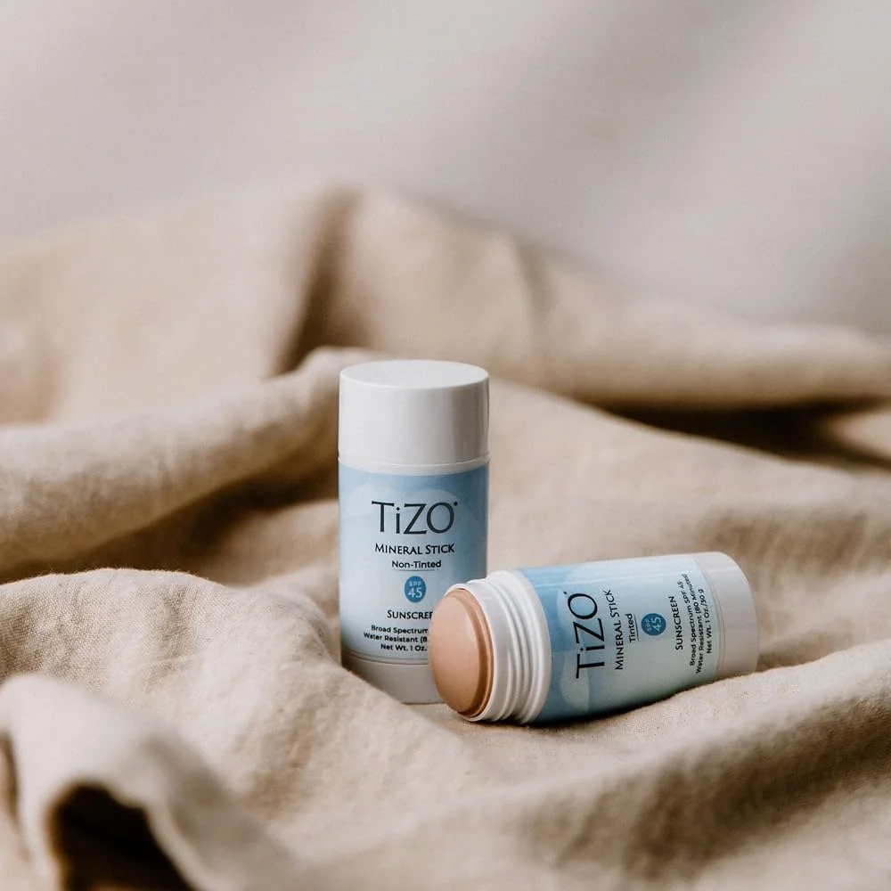 Two Tizo mineral stick sunscreens on a beige fabric surface, one upright and the other lying down with the cap off.