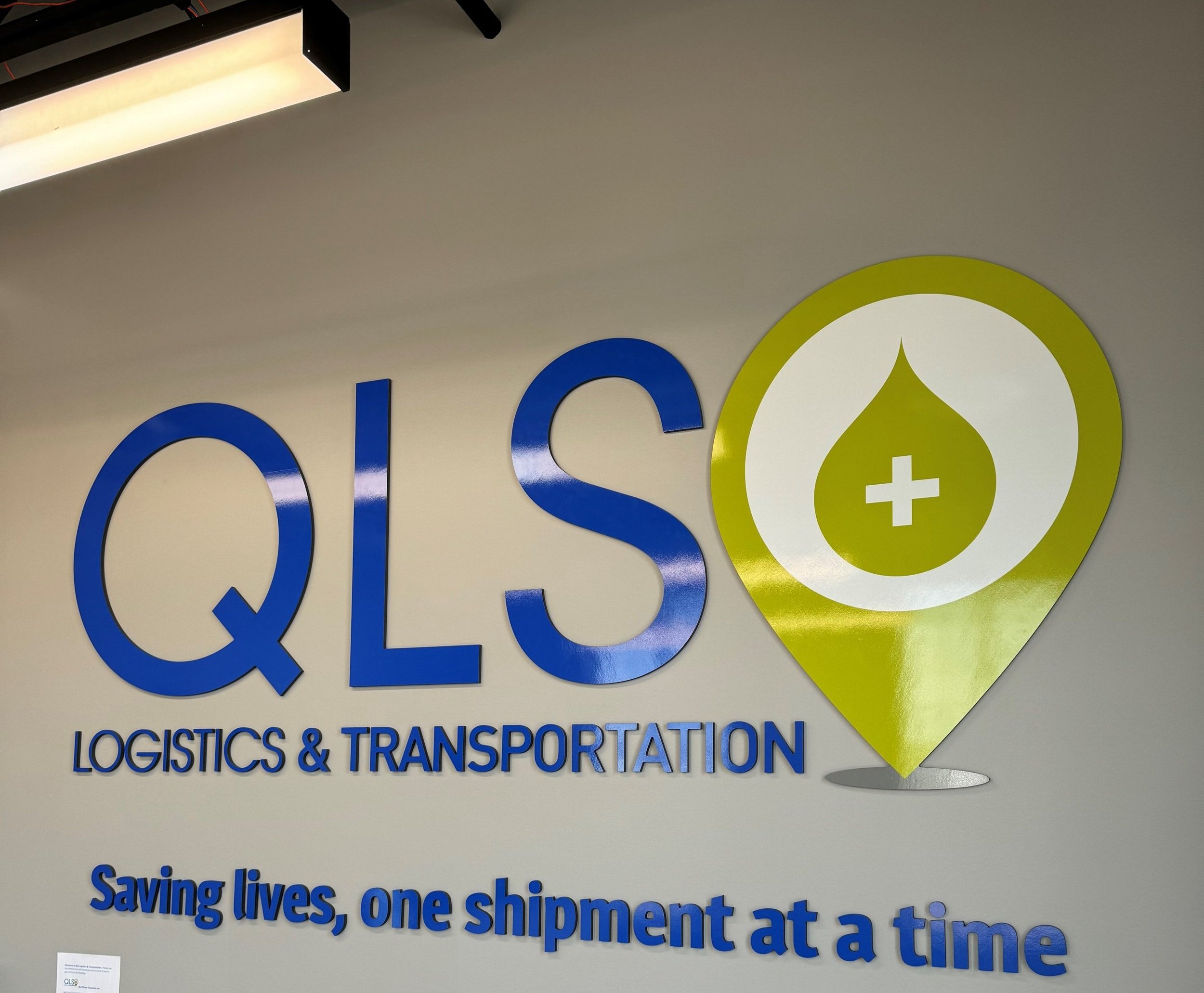 Part of a sign that reads 'Logicistics & Transportation' with a yellow and white logo symbol resembling a droplet with a cross inside. The rest of the sign features blue text on a beige background.