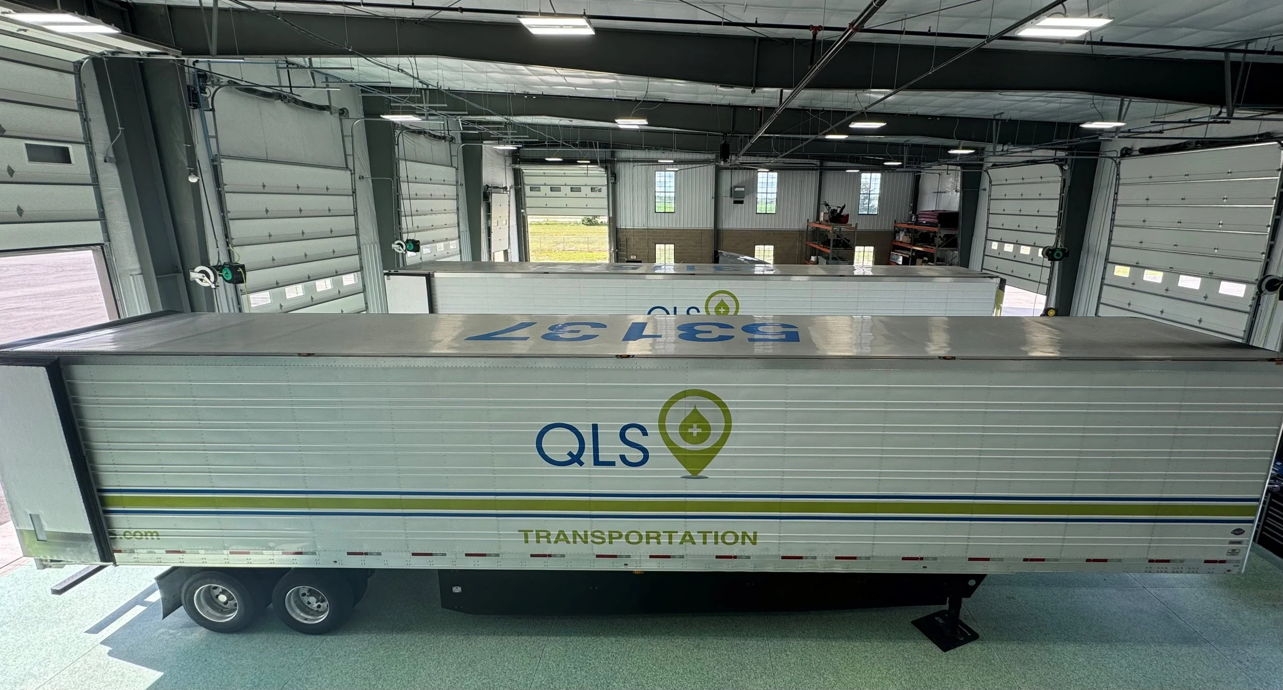 A large white QLS logistics truck parked inside a spacious industrial warehouse with multiple garage doors and windows, visible ceiling pipes, and shelves in the background.
