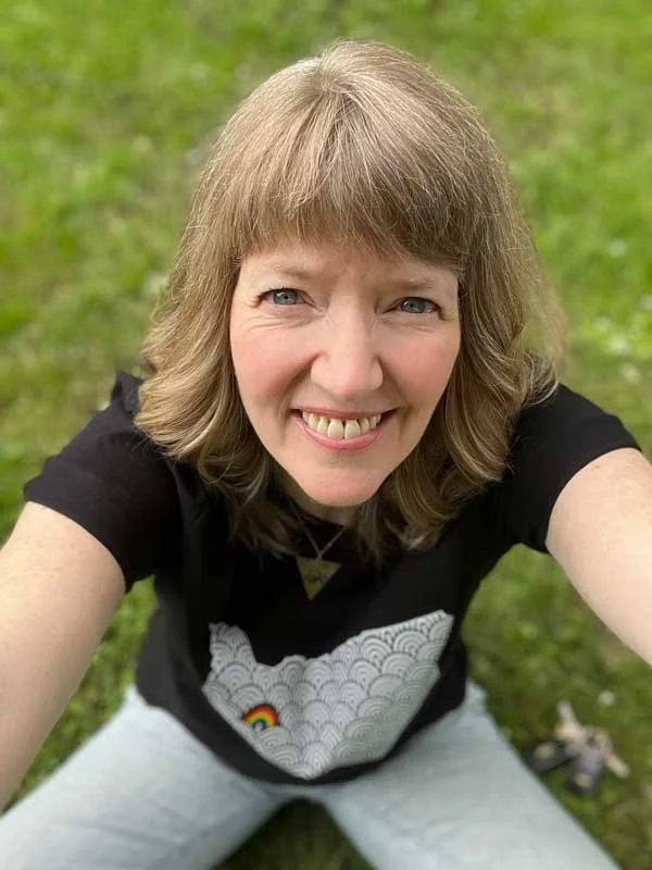 Woman with shoulder-length light brown hair wearing a black T-shirt with a patterned design, smiling up at the camera while sitting on grass.