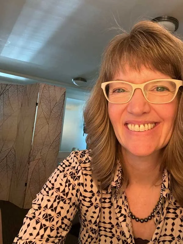 Woman with shoulder-length light brown hair and glasses, smiling indoors while wearing a black and white patterned shirt.