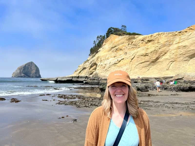 Woman standing on a beach with rocky cliffs and Haystack Rock in the background, wearing a tan hat, blue shirt, and brown cardigan.